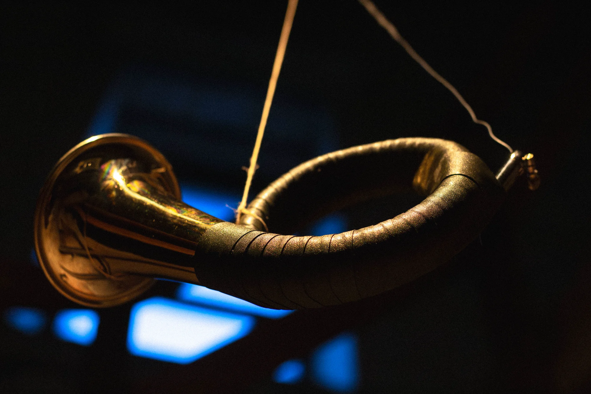 Brass hunting horn suspended by golden wire strings against dark stage, blue light bokeh in background, Hirsch Tot sound installation FEAST & ROADKILL
